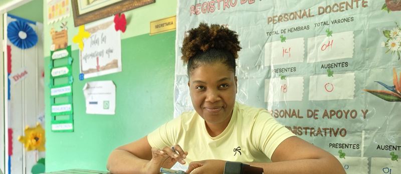 Secretaria de un colegio atendiendo en su escritorio, con el registro de asistencia en la pared